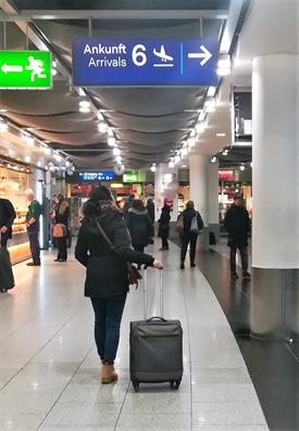 A Young woman dressed in dark clothes and a suitcase is at the Airport. She is Standing below a blue sign which has the word Arrival written on it. She is looking up towards the sign.